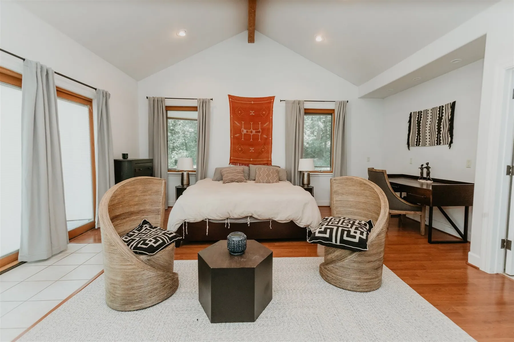 A bedroom with woven armchairs, a low table and a rust-coloured textile above the bed