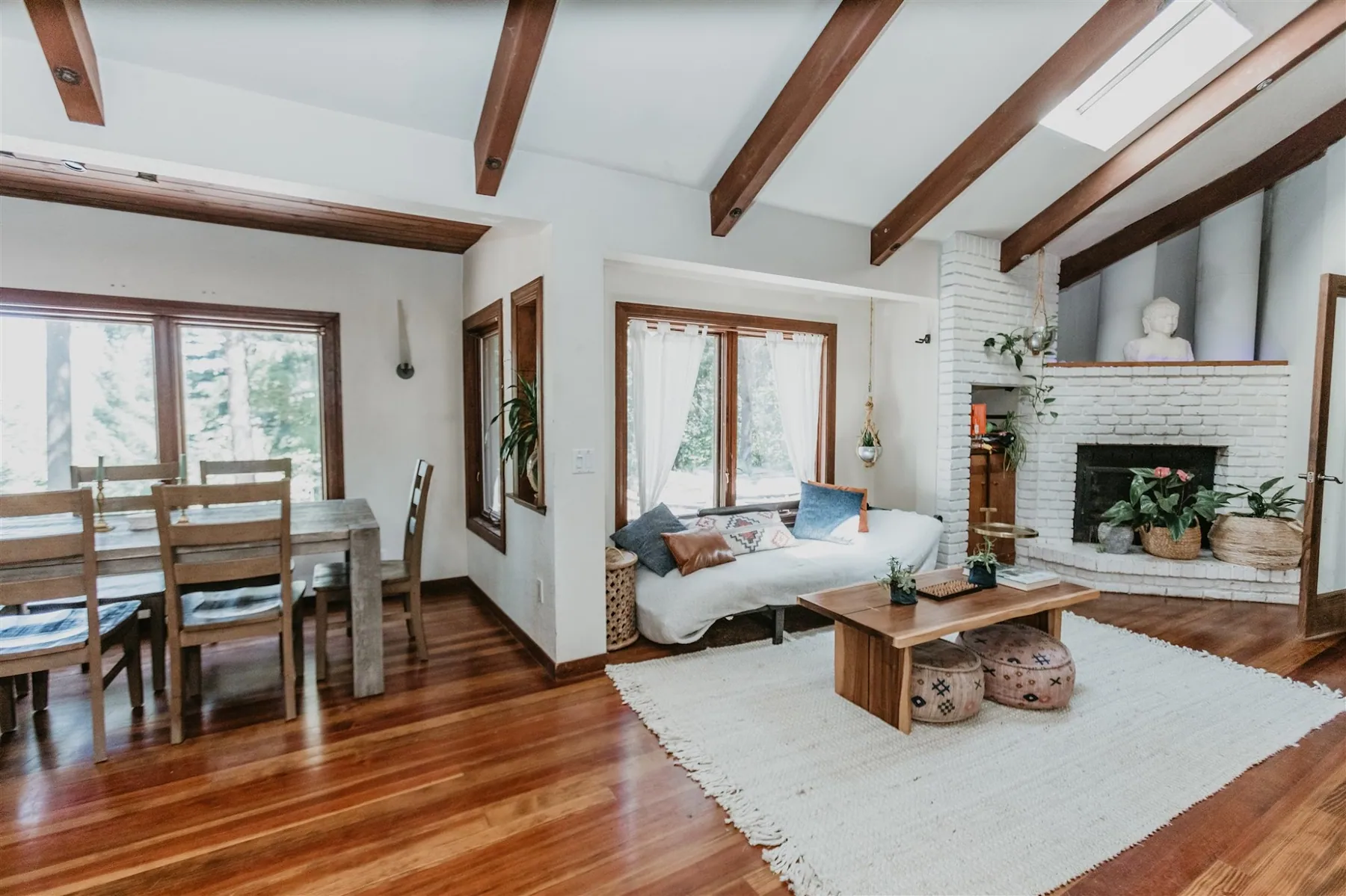 The dining table and living room under exposed beams, with a white brick fireplace