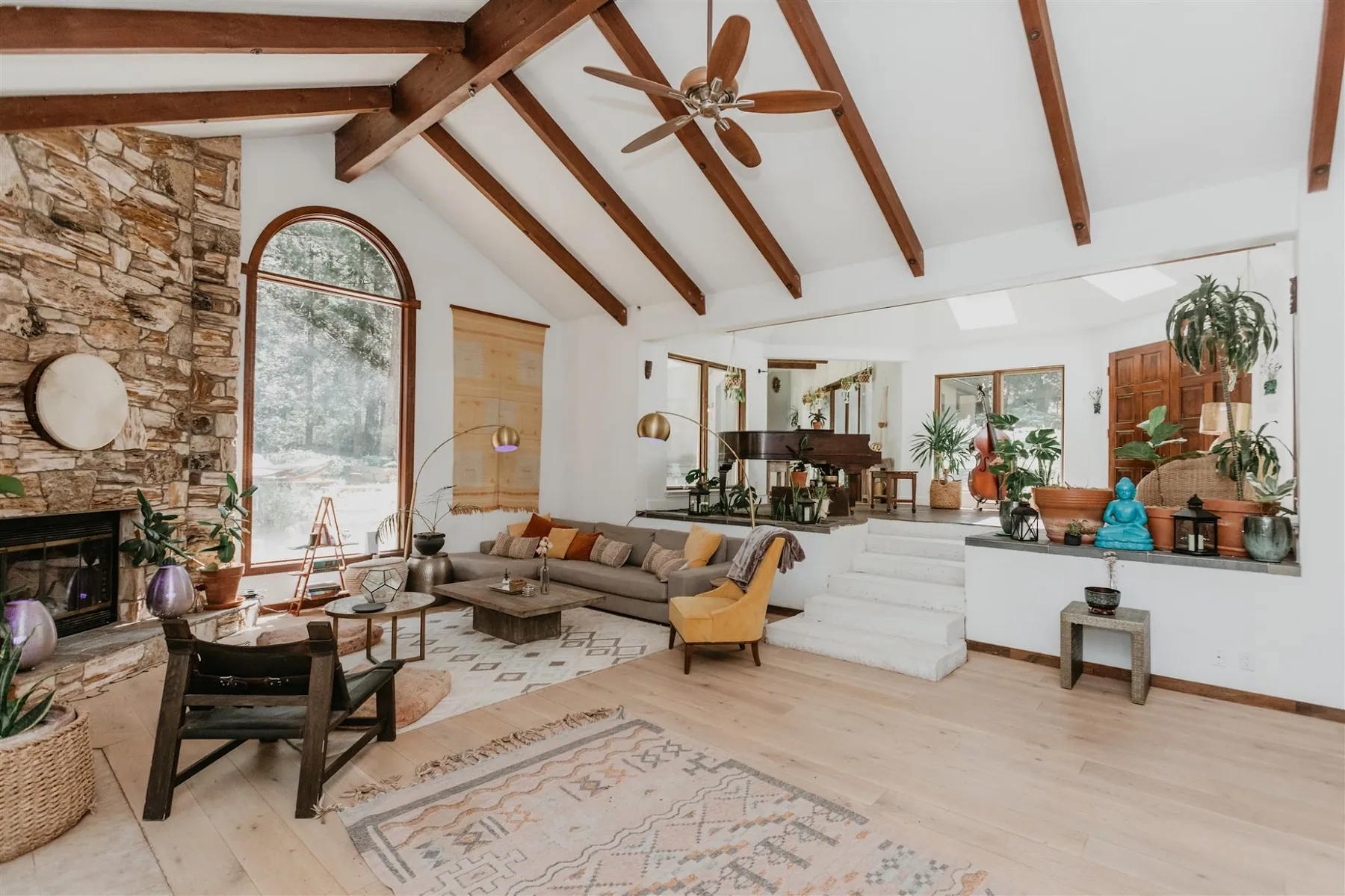The lodge living room with a stone fireplace, vaulted wood beams and an arched window onto the trees