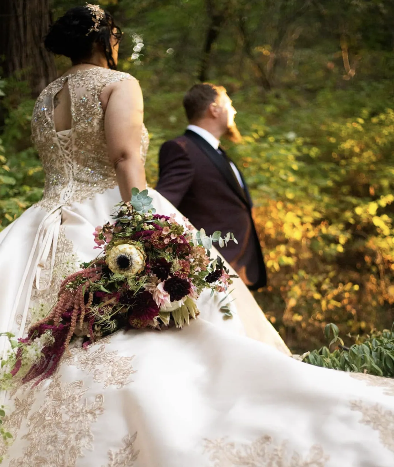 A bride and groom among the trees, her burgundy and cream bouquet held at her side