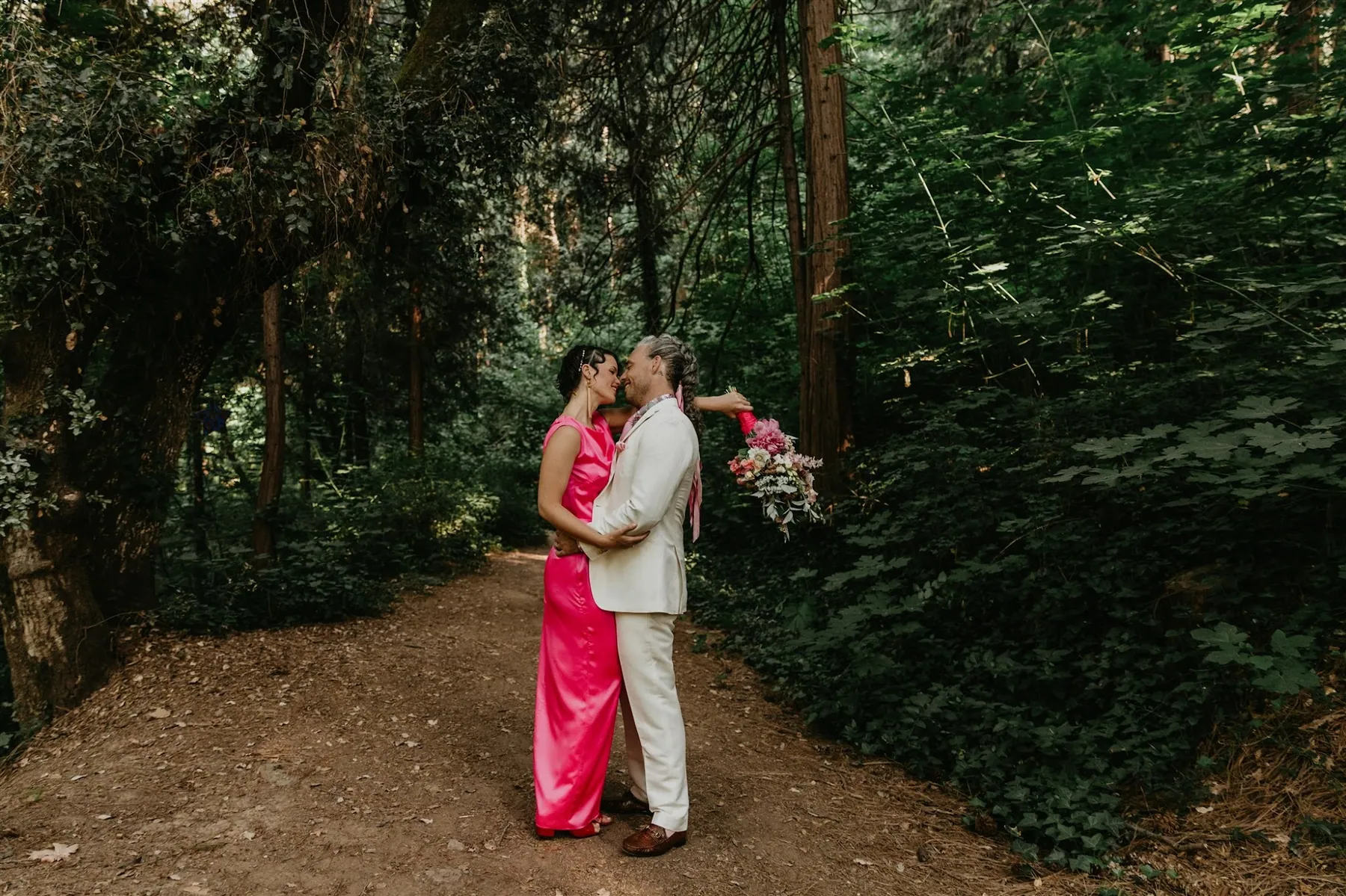A couple embracing on a shaded forest path, the bride in a deep pink gown